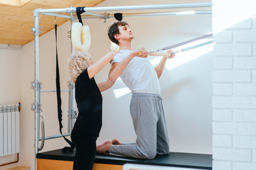 Female blond curly instructor of pilates helping to young man in Cadillac bed.