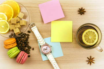 Flat lay. Beautiful ladies watch. Cup of tea with lemon. Stickers for notes, cookies macaroon. Slices of lemon and orange on a saucer. Coffee beans and pieces of sugar on the table