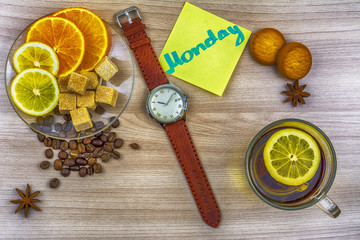 Flat lay. Men's watches with leather strap. Sticker with inscription Monday. Cup of tea with lemon. Slices of lemon and orange on a saucer. Coffee beans, cookies and pieces of sugar on the table