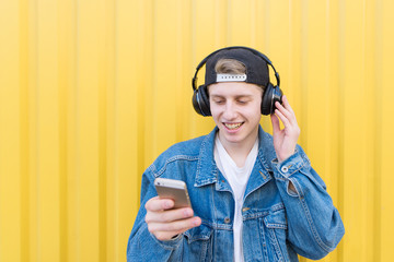 Happy student stands on the background of a yellow wall listening to music in wireless headphones and switching songs on a smartphone. Concept music.