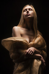 A beautiful young big breasted naked blonde girl, covering her nakedness with a cloth of burlap. Isolated on a black background. Copy space
