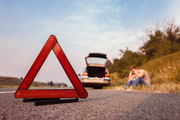 A young man sits on the side of the road. He calls the evacuation service. Warning Triangle in the foreground