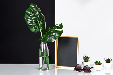 The stylish interior with mock up poster frame, leaves in glass bottle on table with black and white wall on background.