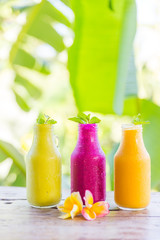 Fresh juice avocado, dragonfruit, mango fruit, healthy drinks in bottles on wooden table with frangipani flowers and babana leafs on background.