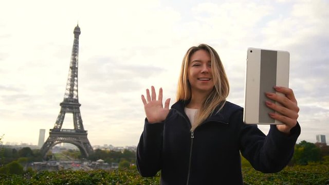 Jocund female person making video call by tablet and front facing camera with Eiffel Tower background in slow motion. Concept of communicating by modern technology and trip to France. Young woman