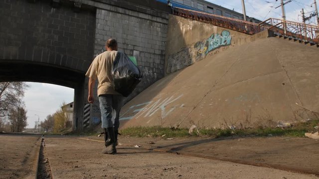 Back view full length of senior homeless male walking to the tunnel under the bridge with trash bin bags on his shoulder. Limping male looking for shelter at sunset. Steadicam stabilized shot.