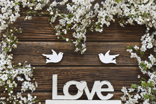 Close-up photo of Beautiful white Flowering Cherry Tree branches with two wooden birds and letters love. Wedding, engagement or betrothal concept on vintage wooden background. Top view, greating card.
