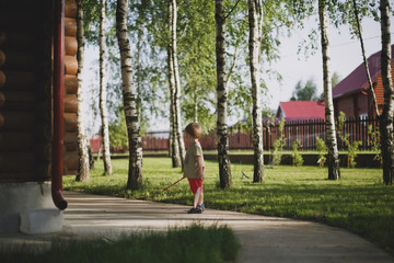 A little blond boy stands with his back next to a country wooden house among birches and holds a badminton racket. Sunny summer day.