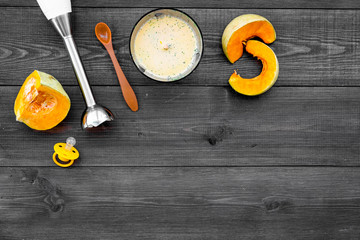 Make baby food at home. Puree with pumpkin near immersion blender and pacifier on dark wooden background top view copy space