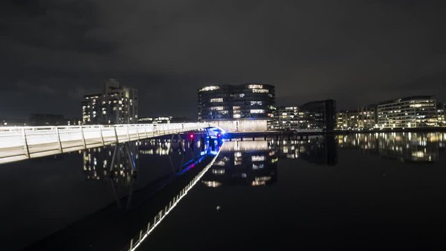 Bike bridge in Copenhagen lighting up in the night and reflecting in the dark quet water, with large office buildings lighting up in the background
