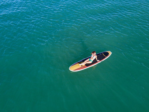 Nondescript fit female in a bikini lounging on her SUP Stand Up Paddle Board in the ocean from a birds eye view