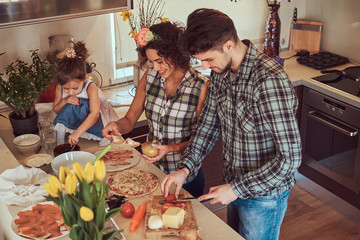 Cute little girl and her beautiful parents prepare pizza while cooking in the kitchen at home.
