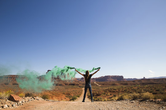Rear view of carefree man with arms raised holding green smoke bombs while standing on dirt road against clear sky amidst desert