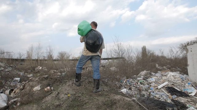 Homeless man with trash bin bags on his shoulder standing on the hill at garbage dump site in city. Slow motion, camera rotating 180 degrees. Environmental problems and pollution concept.