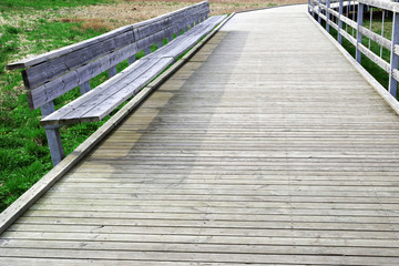 Wide grey brown wooden trail with long bench over green grass.