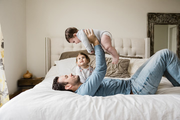 Playful father lifting son while lying by daughter on bed at home