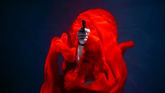 Man is pointing a gun standing underwater behind red fabric.