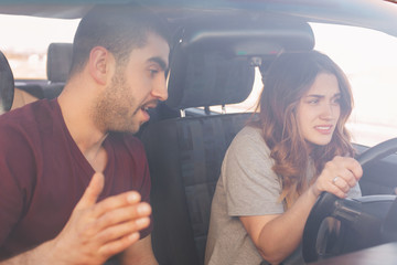 Puzzled young female driver trainee drives car, listens attentively male instructor who gives some...