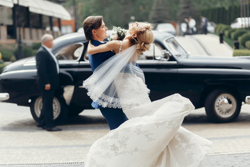 Romantic bride and groom portrait after wedding ceremony, happy newlywed couple dancing outdoors, luxury car in background, happy groom hugging bride