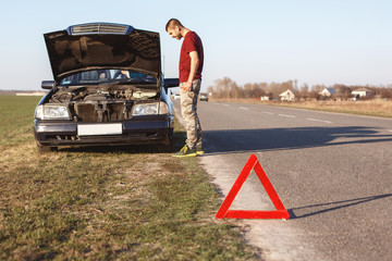 Outdoor shot of male stands near his brocken car, can`t solve problem by himself red triangle as...