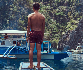 Shirtless Asian guy standing on a bow of the ship on the background of a beautiful landscape of rocky bay.