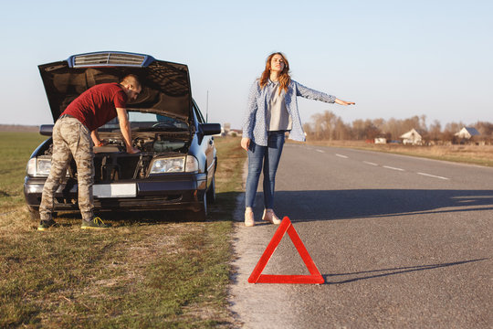 Desperate female stops vehicles on road as needs help, travel together with husband by car and have damage with engine, can`t solve problem by themselves, red warning triangle in foreground.