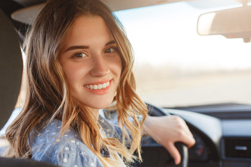 Back view of pretty smiling woman with broad smile, has attractive look, sits at wheel in car, has break after long trip, looks directly into camera. Positive woman drives automobile, enjoys speed