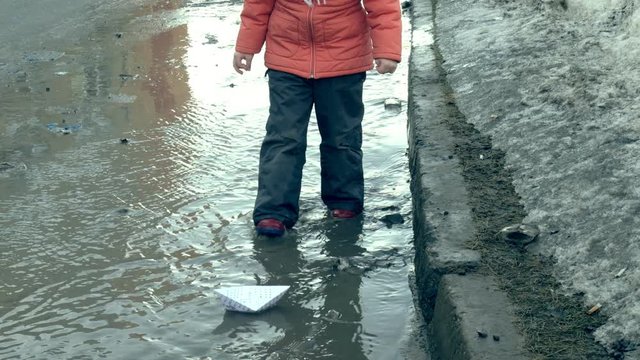 Girl in rubber boots jumping on puddle near wite paper boat