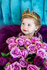 Portrait of little princess baby in purple dress and diadem on blue sofa with bouquets of roses