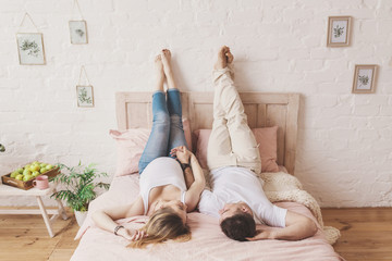 Young future parents in bright clothes lie next to each other on a large bed in a large cozy bedroom and raise their feet up against a white wall
