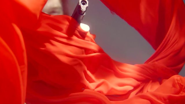 Close up shot of a man holding a firearm underwater behind falling red dress.