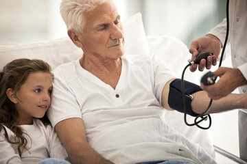 Elderly man receiving a hospital visit from his granddaughter while having his blood pressure taken.