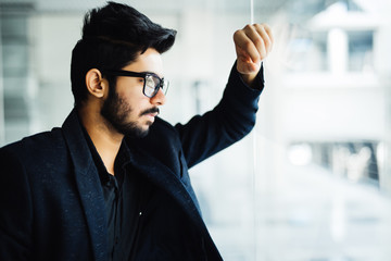 Indian happy young business man in eyeglasses work in modern office looking thought window