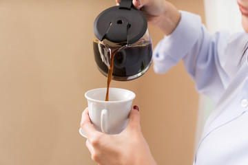 Close up of pouring hot coffee from glass container into white ceramic cup by woman' s hand. Beautiful young woman in her room pouring herself a mug of hot filtered coffee from glass pot. - Powered by Adobe