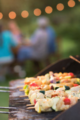 close-up of a barbecue where chicken skewers are cooking, friends gathered around a table to share a meal.
