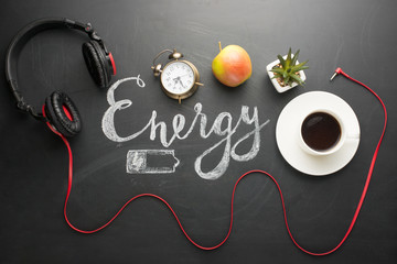 A white cup of coffee on a dark texture with an inscription of energy and a battery and an old alarm clock an apple and red headphones The concept of an energetic morning, and the beginning of the day