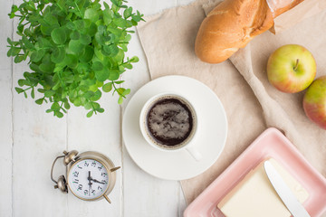 Morning cup of coffee, alarm clock, apples, butter and baguette, in a light kitchen. Background area, the concept of a bright morning and breakfast. View from above
