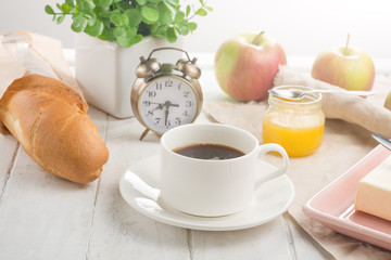 Morning cup of coffee, alarm clock, apples, butter and baguette, in a light kitchen. Background area, the concept of a bright morning and breakfast