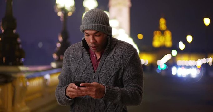 Modern black man vacationing in Paris using his smartphone on a nippy night outside, Cold young man texting his friend via smartphone on the Pont Alexandre III, 4k
