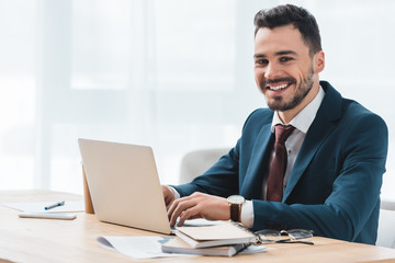 handsome young businessman using laptop and smiling at camera in office