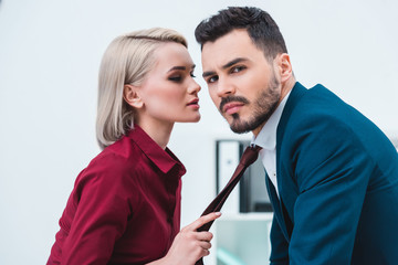 young businesswoman holding necktie of handsome businessman looking at camera in office