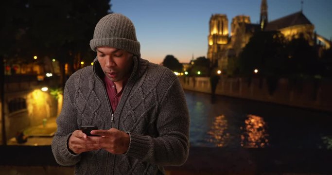 Young man vacationing in Paris communicating with someone on his smartphone via texting on a chilly evening, African male trying to reach a friend on his phone with Notre Dame in the background, 4k
