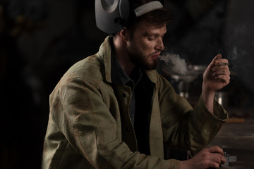 workman with lifted up protective mask sitting and smoking cigarette at factory