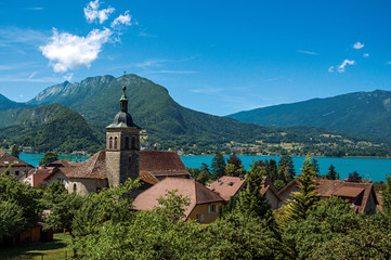 Fototapeta na wymiar View of houses and belfry with blue sky mountains landscape on background, in the village of Talloires. A lovely village next to the Lake of Annecy. Department of Haute-Savoie, southeastern France.