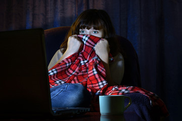 Scared young woman looks terrible movie at night on laptop