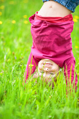 Happy little girl standing upside down on green grass in summer park