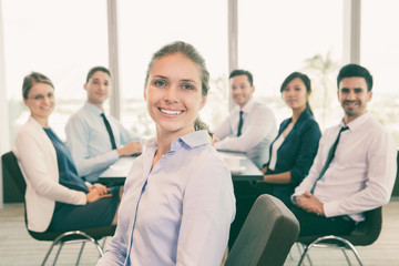 Obraz premium Portrait of happy female executive manager sitting on office chair, looking at camera and smiling. Her business team on background
