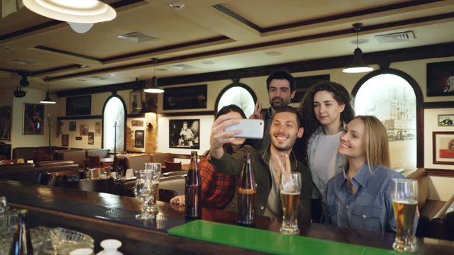 Friends are taking selfie with smartphone in bar. Young people are posing, laughing and talking. Beer bottles and glasses in foreground