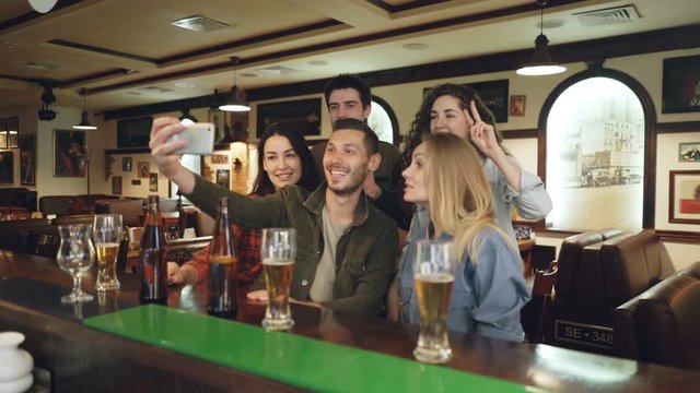 Young people are using smartphone to take selfie in popular bar. Friends are posing, having fun, gesturing and laughing.