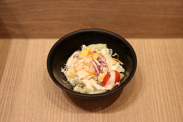 Vegetable salad in the small black bowl on the wooden floor. It is a dish of various mixtures of raw vegetables.